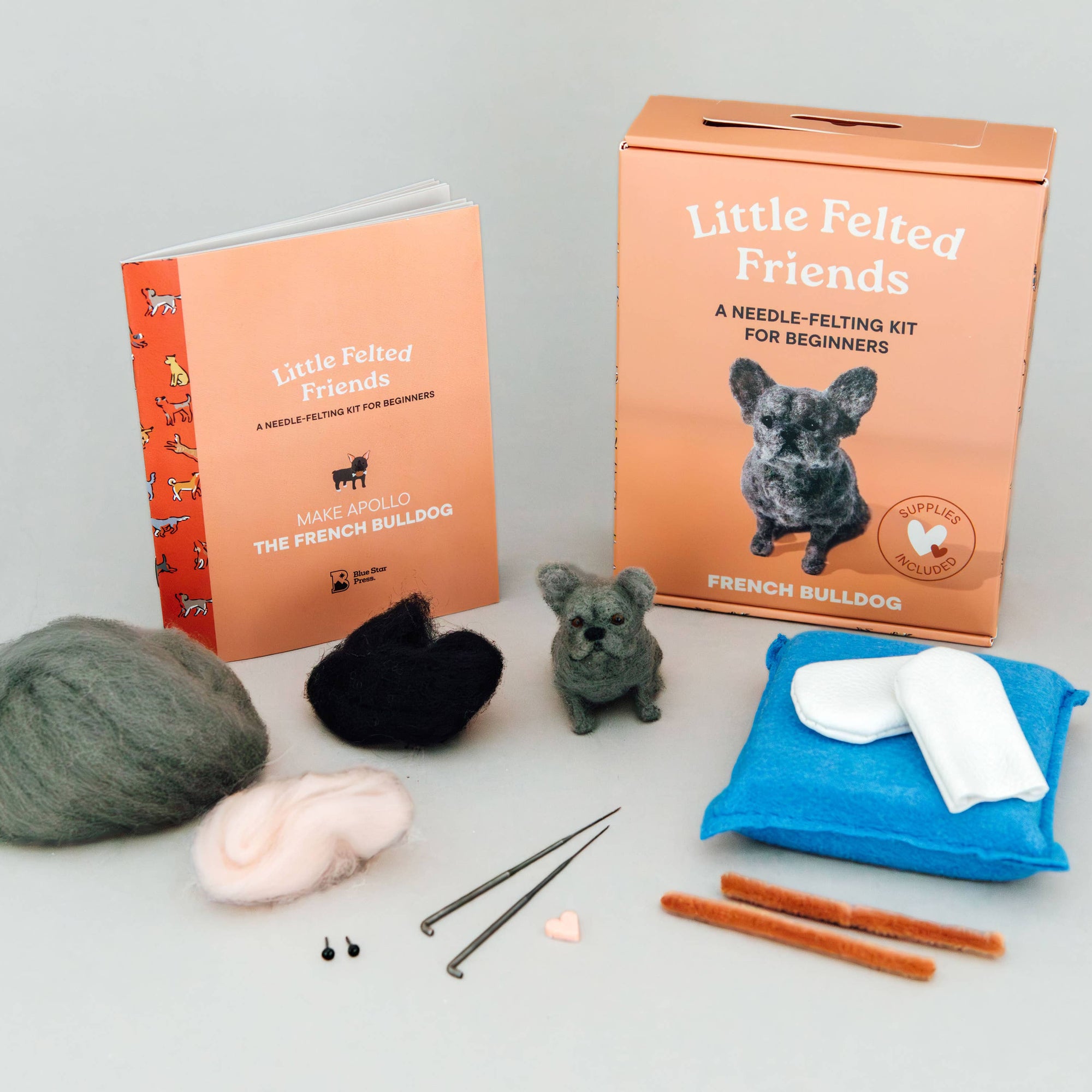 A needle felting kit for beginners titled 'Little Felted Friends' displayed on a light gray background, including an orange box, a booklet, wool balls, a black felt piece, a small dog figurine, a blue padded mat with white stuffing, metal needles, and wooden tools.