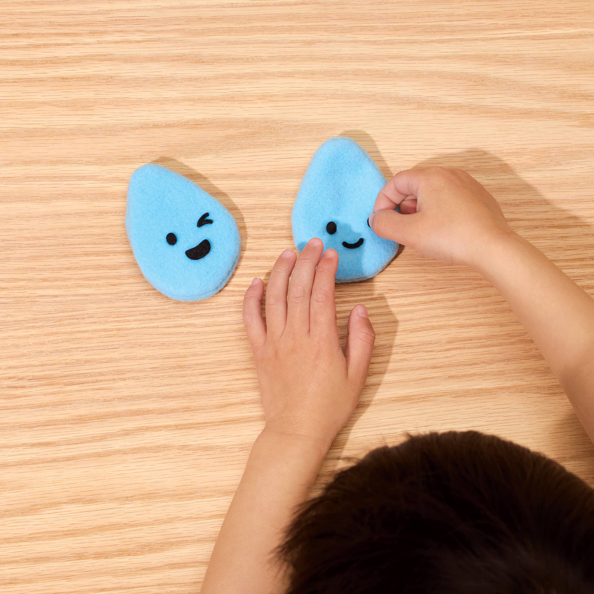 Two small blue teardrop-shaped plush toys with embroidered faces on a light wood tabletop, with a child's hands and dark hair visible in the foreground.