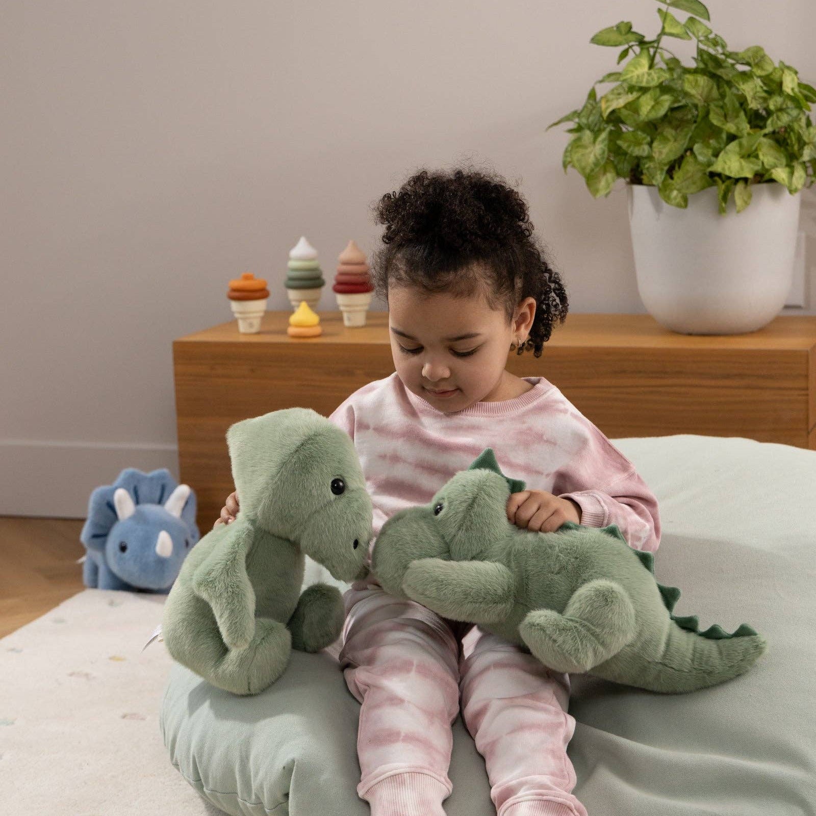 Child playing with green dinosaur plush toys on a bean bag in a room with a plant and wooden shelf.