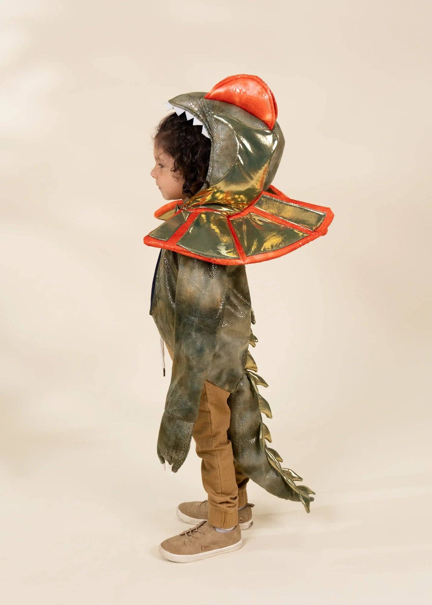 A child wearing a dinosaur costume, featuring a green and brown textured body, a red hood with white teeth, and a red-trimmed collar, standing against a plain beige background.