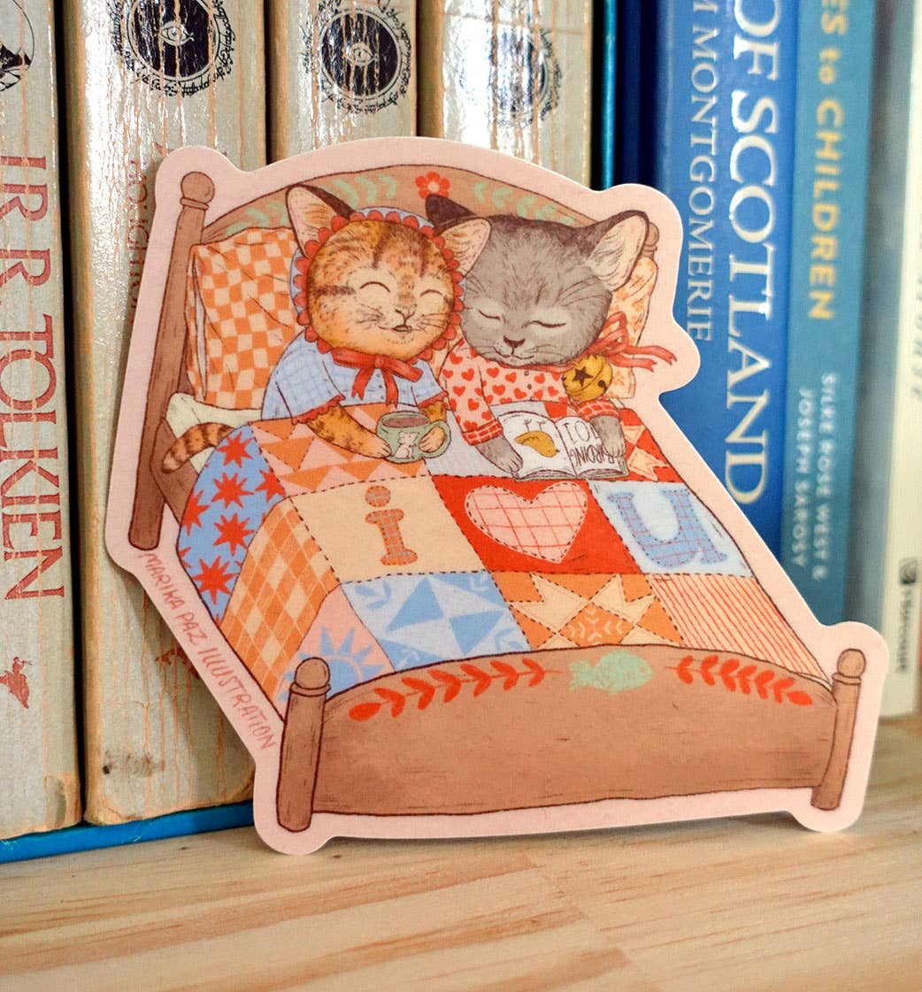 Illustrated sticker of two cats in bed with a quilt, placed on a wooden surface with books in the background.