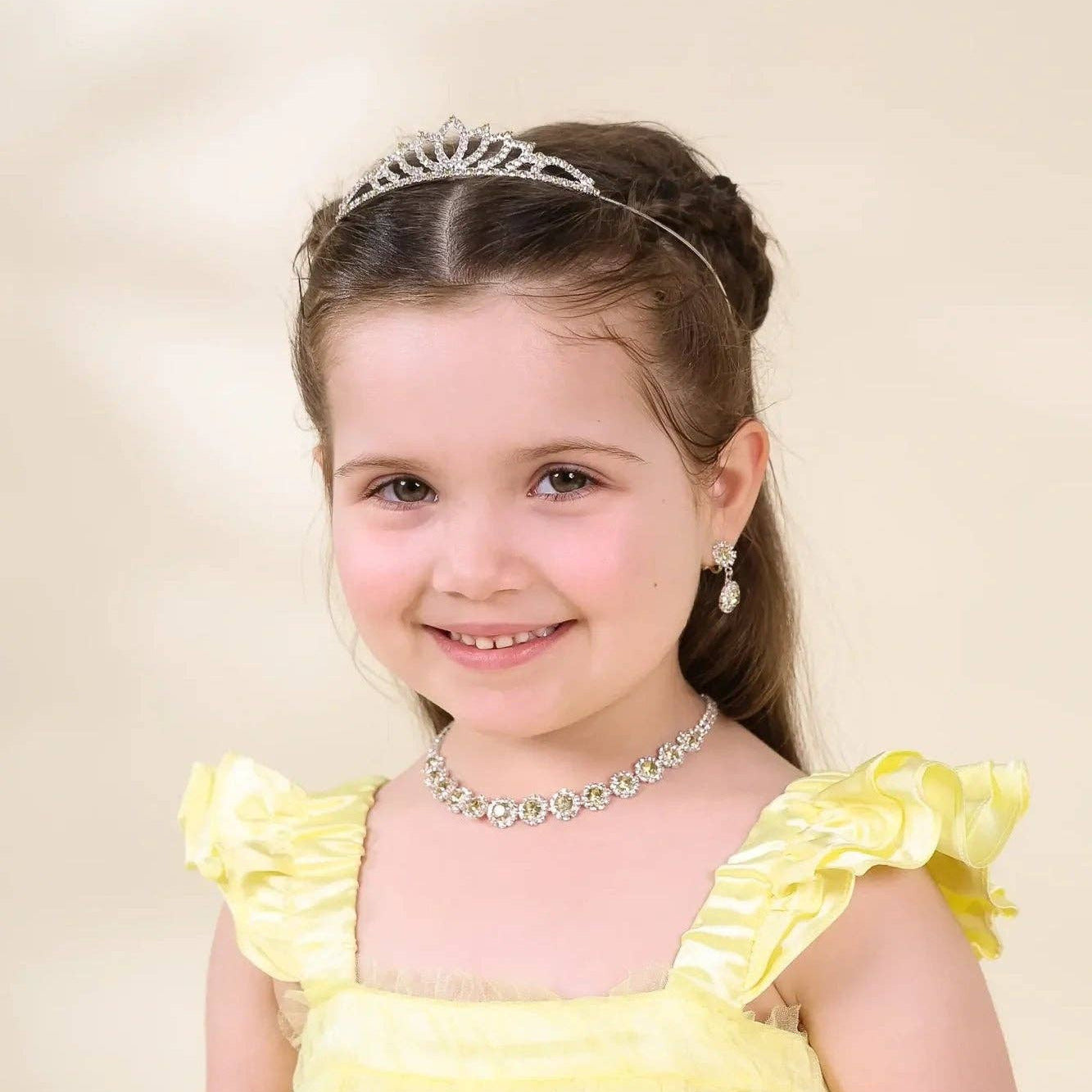 A young girl with brown hair in a half-up style wearing a silver tiara, matching silver necklace, and a yellow dress with ruffled shoulders, photographed against a soft beige studio background.
