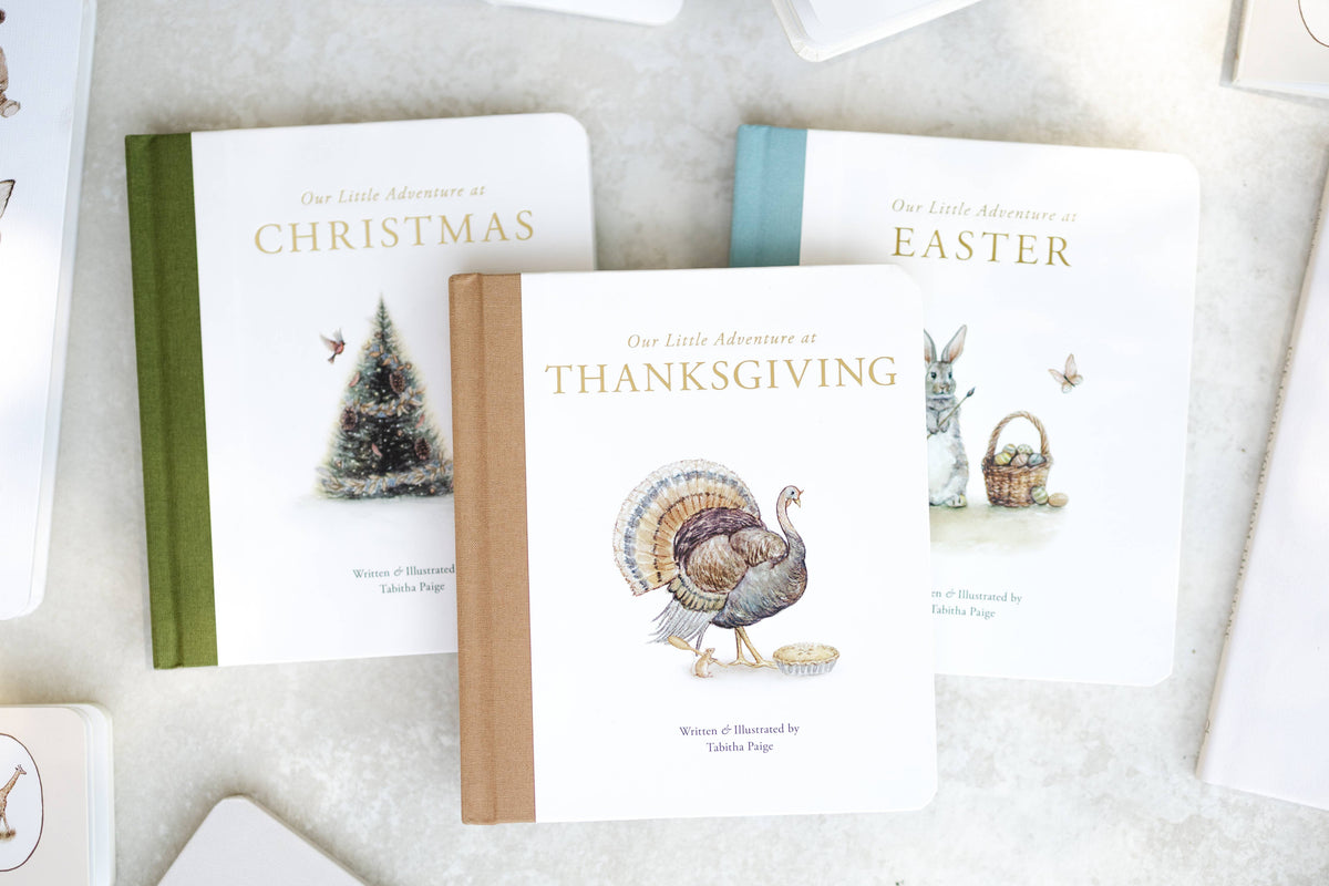 Three children's picture books titled 'Our Little Adventure at Thanksgiving,' 'Our Little Adventure at Christmas,' and 'Our Little Adventure at Easter' arranged on a light textured surface. The Thanksgiving book is centered in front, showing a watercolor turkey illustration on a white cover with a tan spine, while the other two books are partially visible behind it.