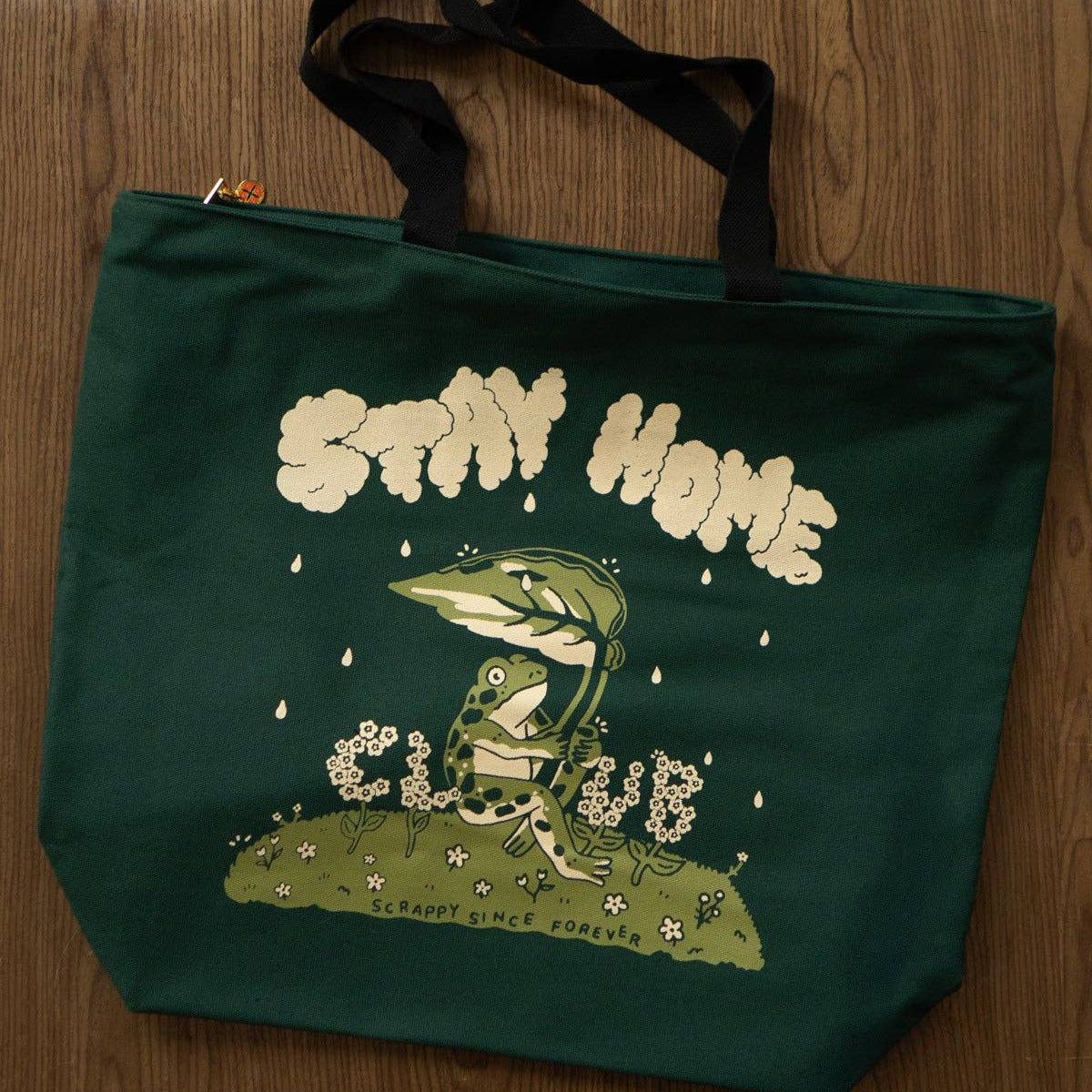 A dark green canvas tote bag with black handles, featuring a graphic of a green frog sitting in a flower under the words 'STAY HOME' and 'CLUB', placed on a wooden surface with books in the background.