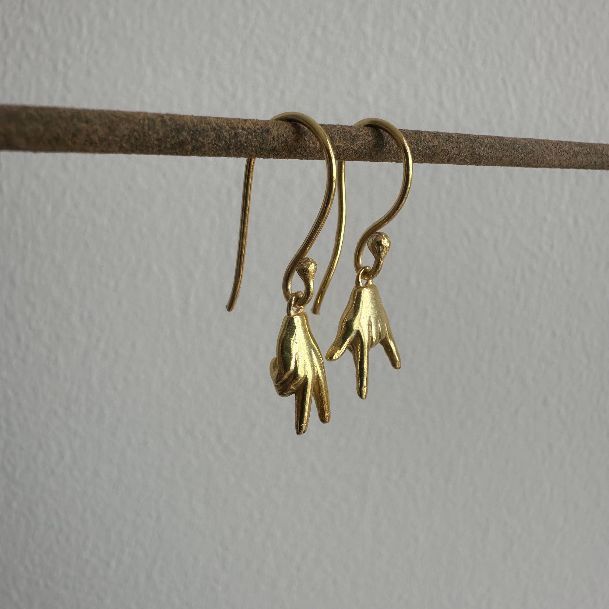 Gold hand-shaped earrings hanging on a thin brown rod against a light gray background