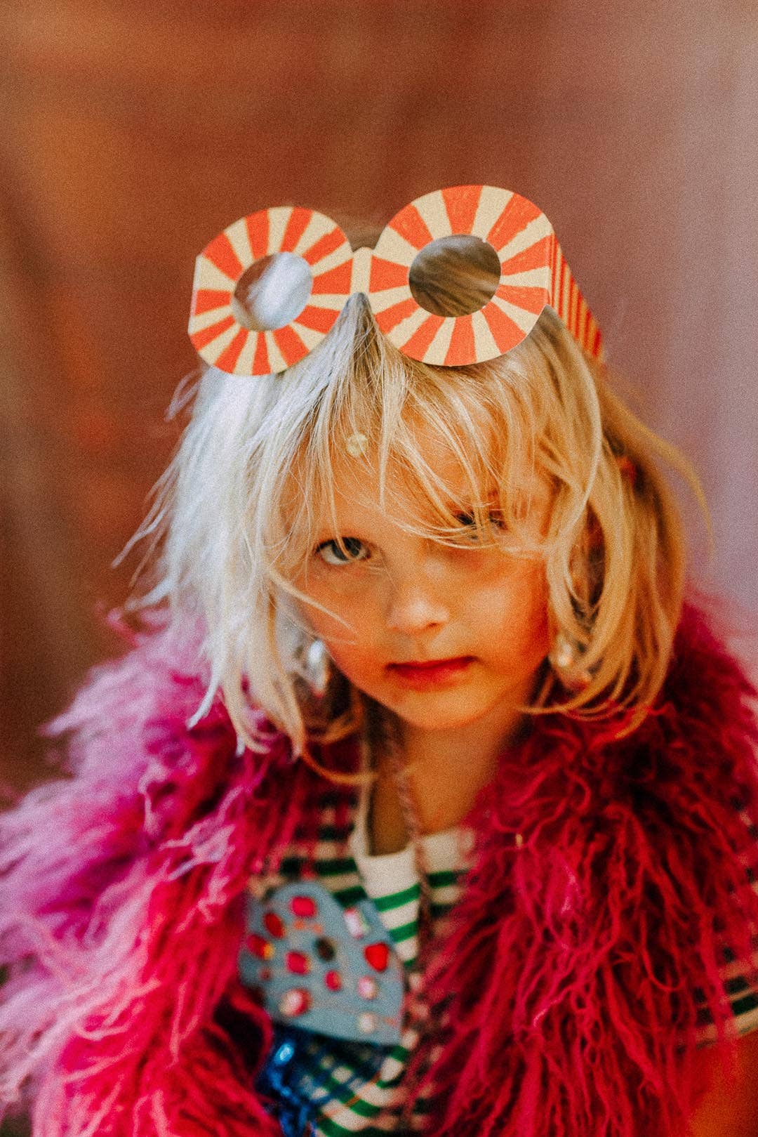 A young child wearing novelty round sunglasses with red and white diagonal stripes, styled with a bright pink fluffy scarf and a striped shirt.