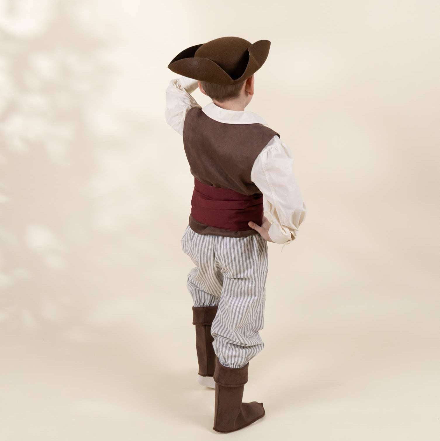 A child wearing a pirate costume, photographed from behind against a warm beige studio background with soft leaf shadows. The costume includes a brown hat, brown vest, white shirt, dark red waist cloth, striped pants, and brown boots.