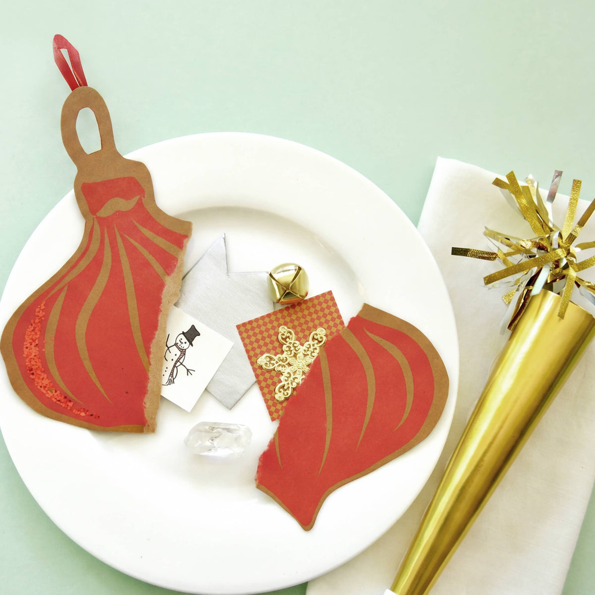A styled flat-lay of two red and gold paper dress-shaped ornaments on a white plate, alongside a gold party popper, a napkin, and small decorative cards on a pale mint green background.