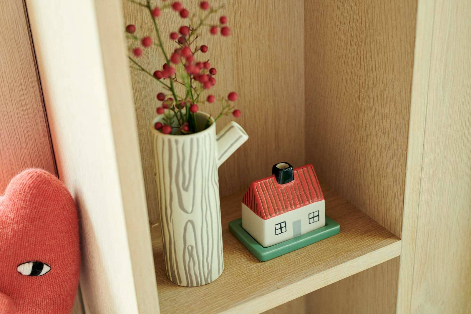 A ceramic wood-grain pitcher vase holding red berries, displayed on a light wood shelf next to a small house-shaped ceramic container.