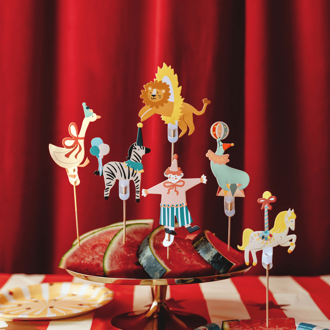A set of colorful circus-themed paper party toppers on sticks, including a lion, zebra, giraffe, clown, and carousel horse, displayed on a gold stand with watermelon slices in front of a red curtain backdrop.