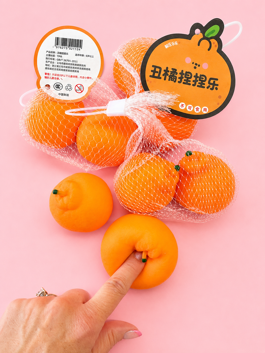 Three orange-shaped novelty squishy toys with mesh packaging and a cartoon tag, displayed on a pink background with a hand pressing down on one toy.