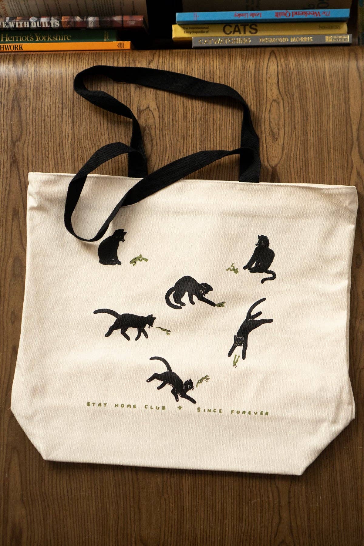 An off-white canvas tote bag with black handles, featuring black cat silhouettes and the text 'STAY HOME CLUB SINCE FOREVER' printed on the front, displayed on a wooden surface.