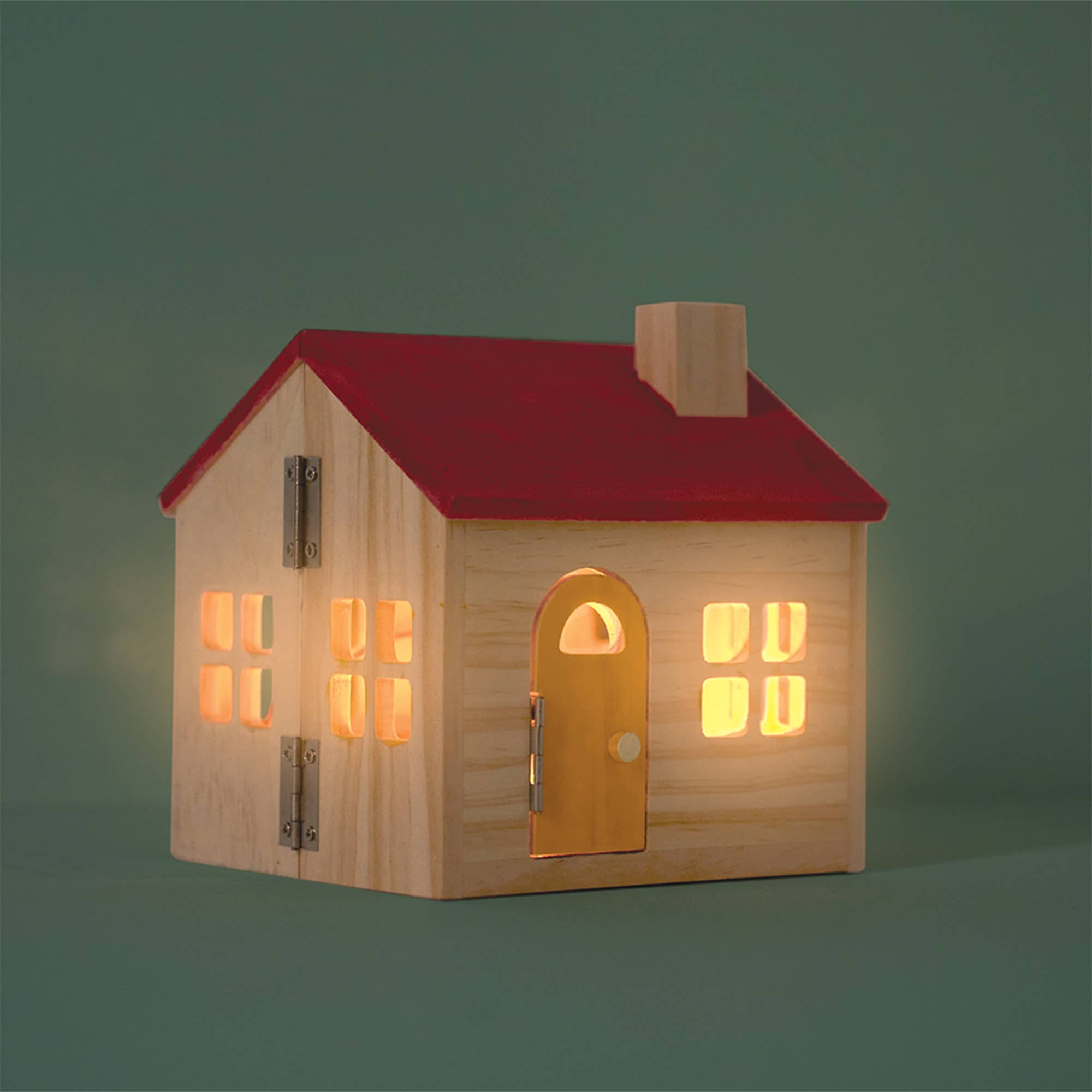 A miniature wooden house figurine with a red roof, natural wood walls, and glowing warm yellow lights in the windows, set against a solid dark green background.