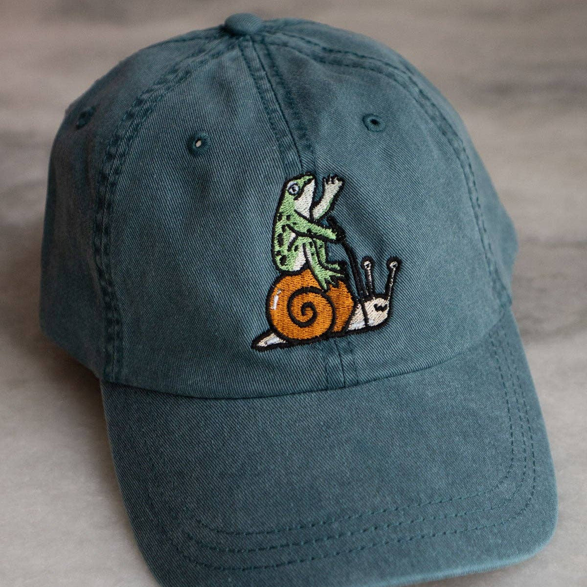 Blue cap with embroidered design of a frog on a snail, on a light wooden surface.