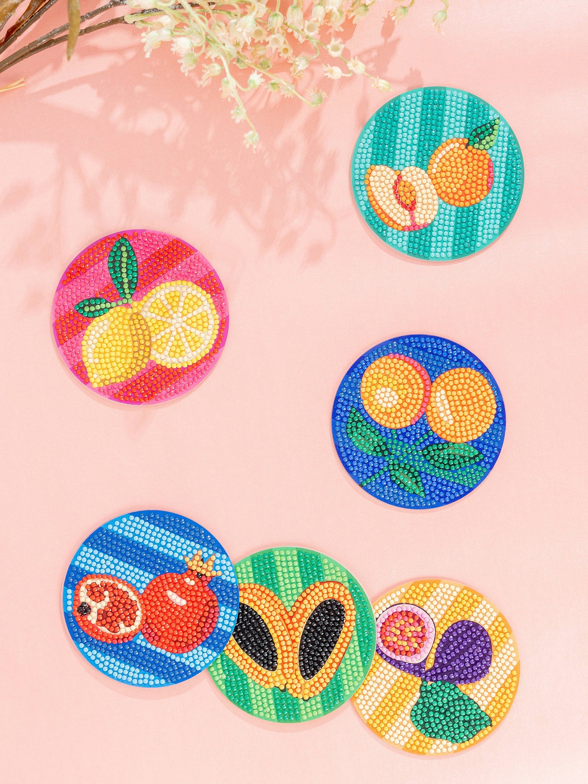 Colorful beaded fruit coasters on a pink surface with a decorative plant.