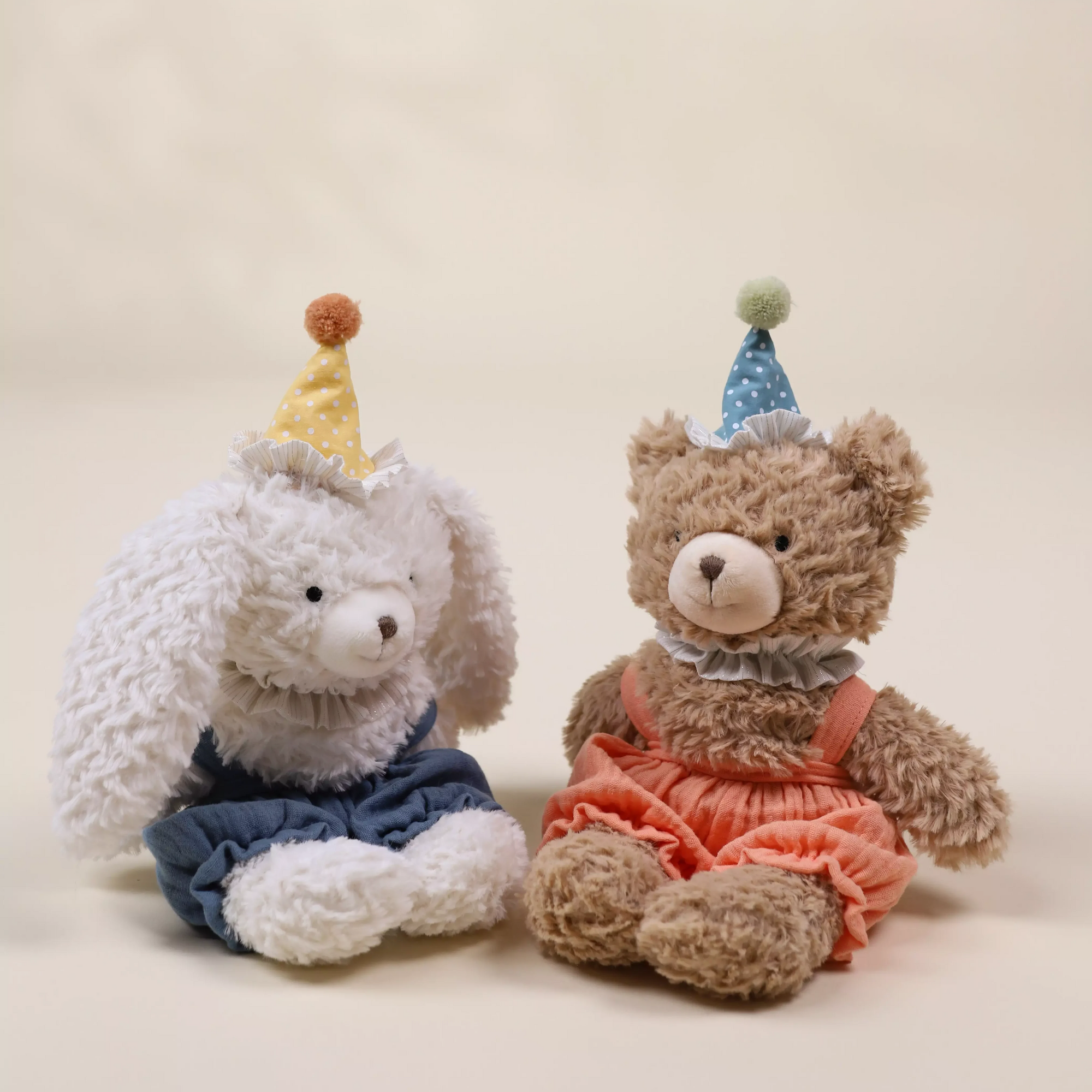 Two stuffed animals, one resembling a bunny and the other a bear, wearing party hats and dresses on a plain background.