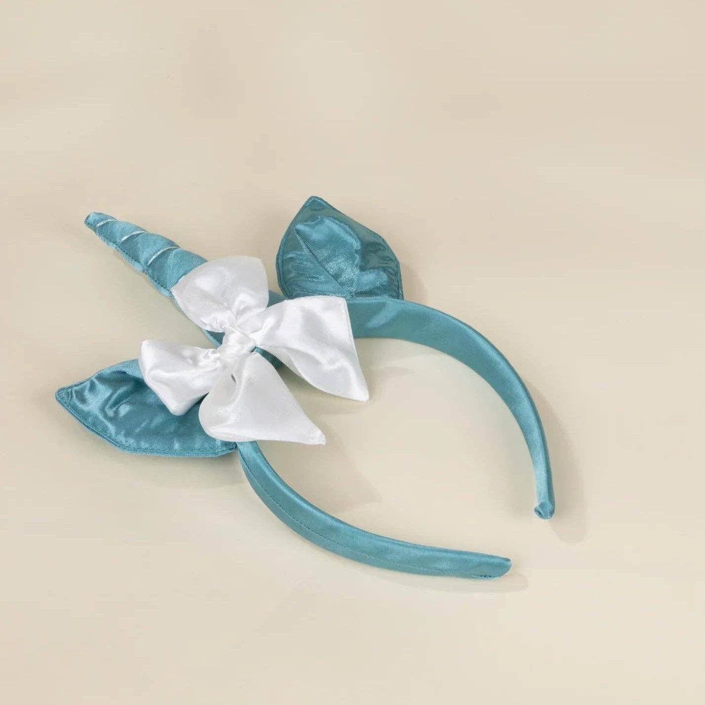 A satin unicorn headband in a dusty teal blue color, featuring a horn, a bow, and a leaf accent, displayed on a solid beige background.