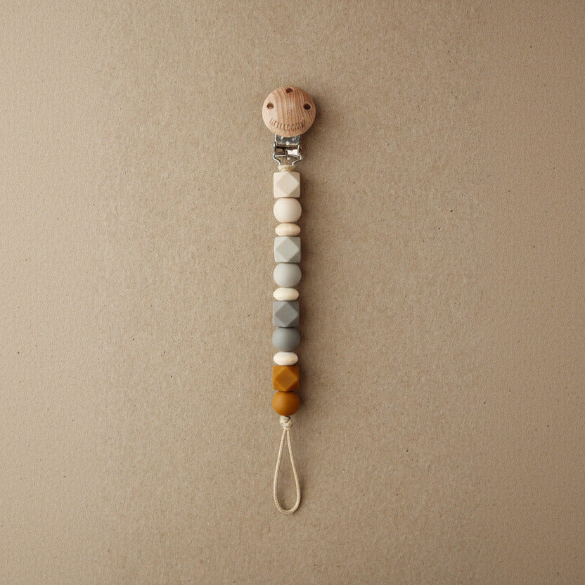 A wooden baby pacifier clip with colorful silicone beads in shades of gray, cream, and mustard yellow, shown on a textured beige background.
