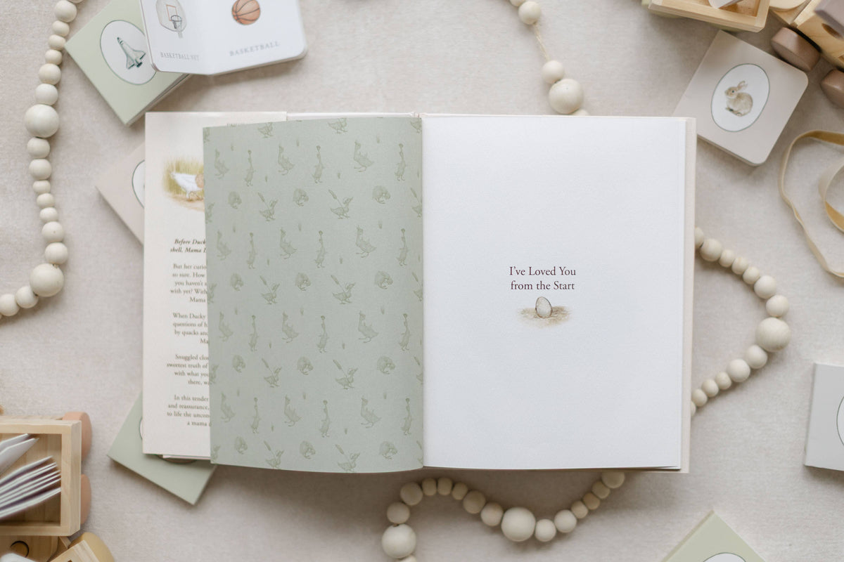 An open baby keepsake book laid flat on a neutral fabric surface, surrounded by wooden beads, small cards, and baby accessories. The left page has a pale sage green botanical pattern, and the right page is white with the printed text 'I've Loved You from the Start' and a small illustration.