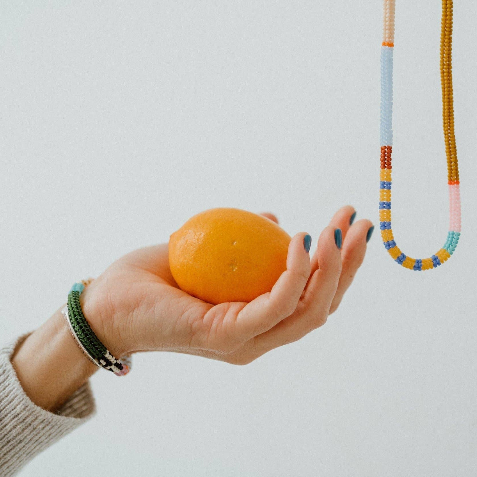 Hand holding an orange with a colorful beaded necklace on a plain background