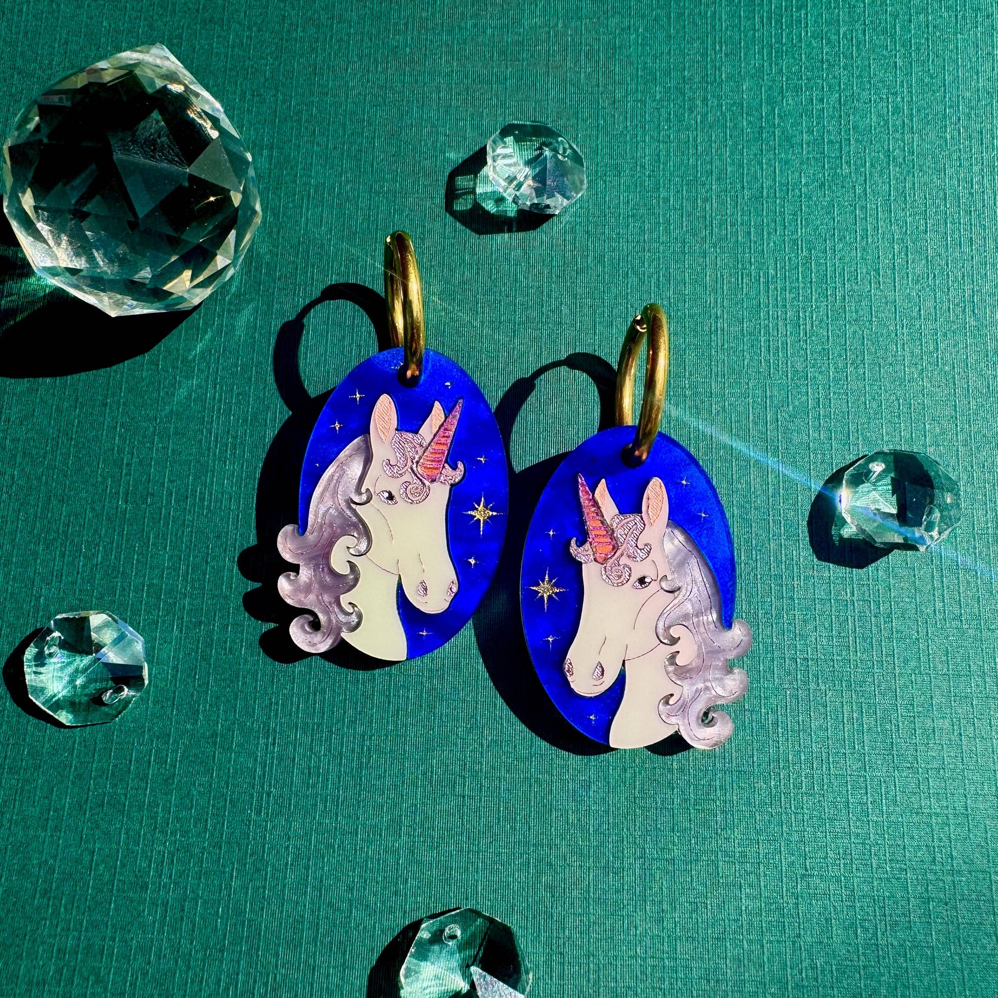 Unicorn-themed earrings on a teal background with crystal-like decorations.