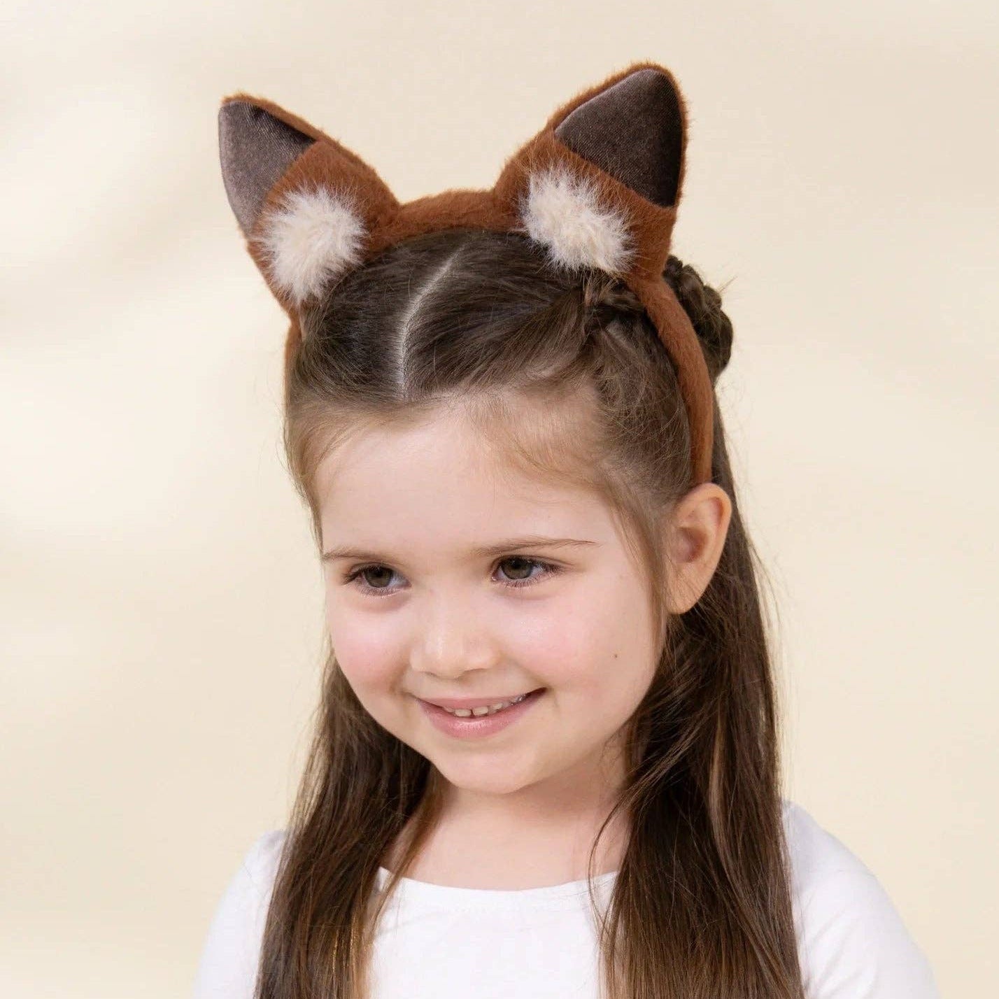 A young girl with long brown hair wearing a brown and white faux fur animal ears headband, posing against a solid light beige background.