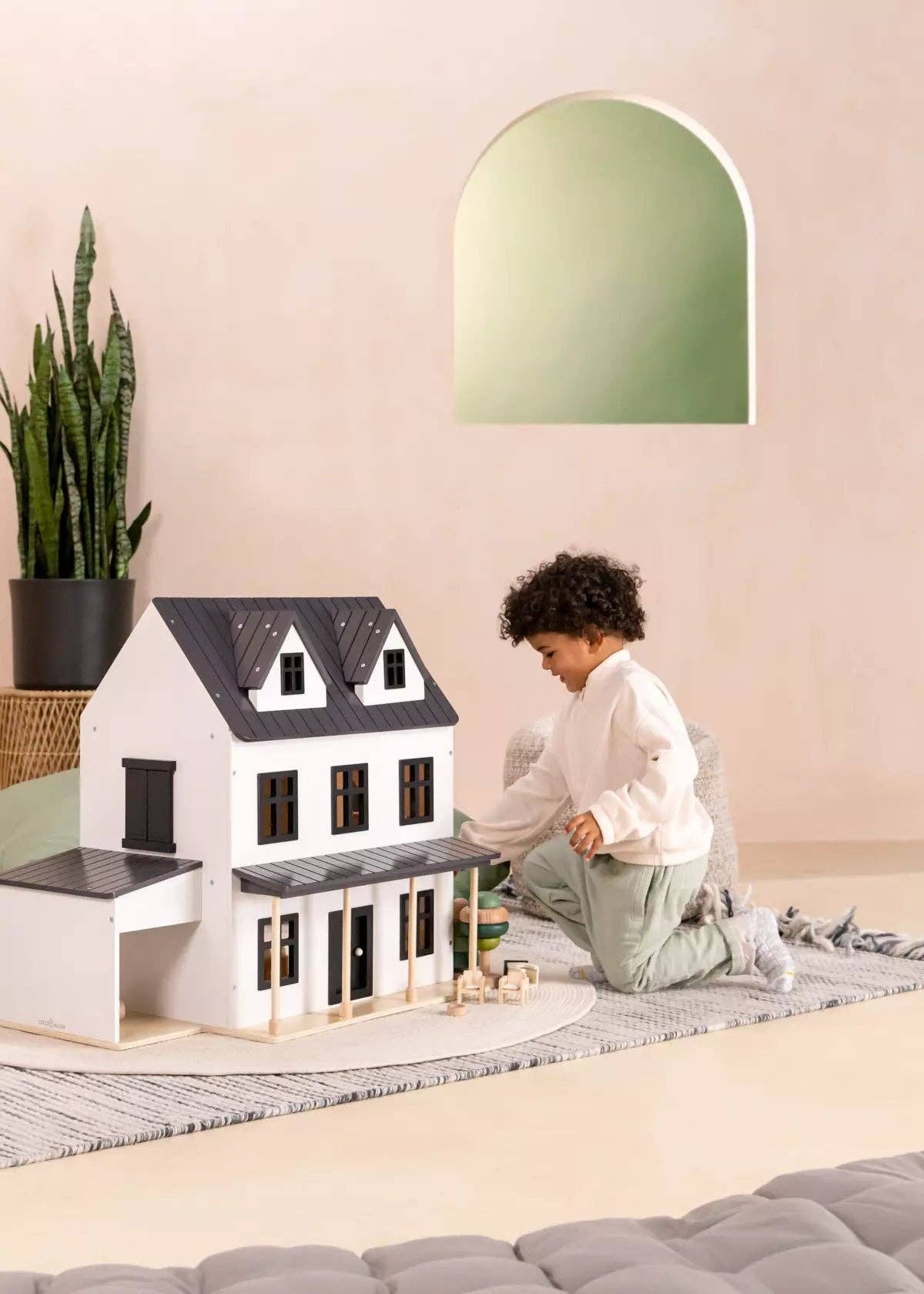 Child playing with a dollhouse on a rug in a room with a green lamp and plant.