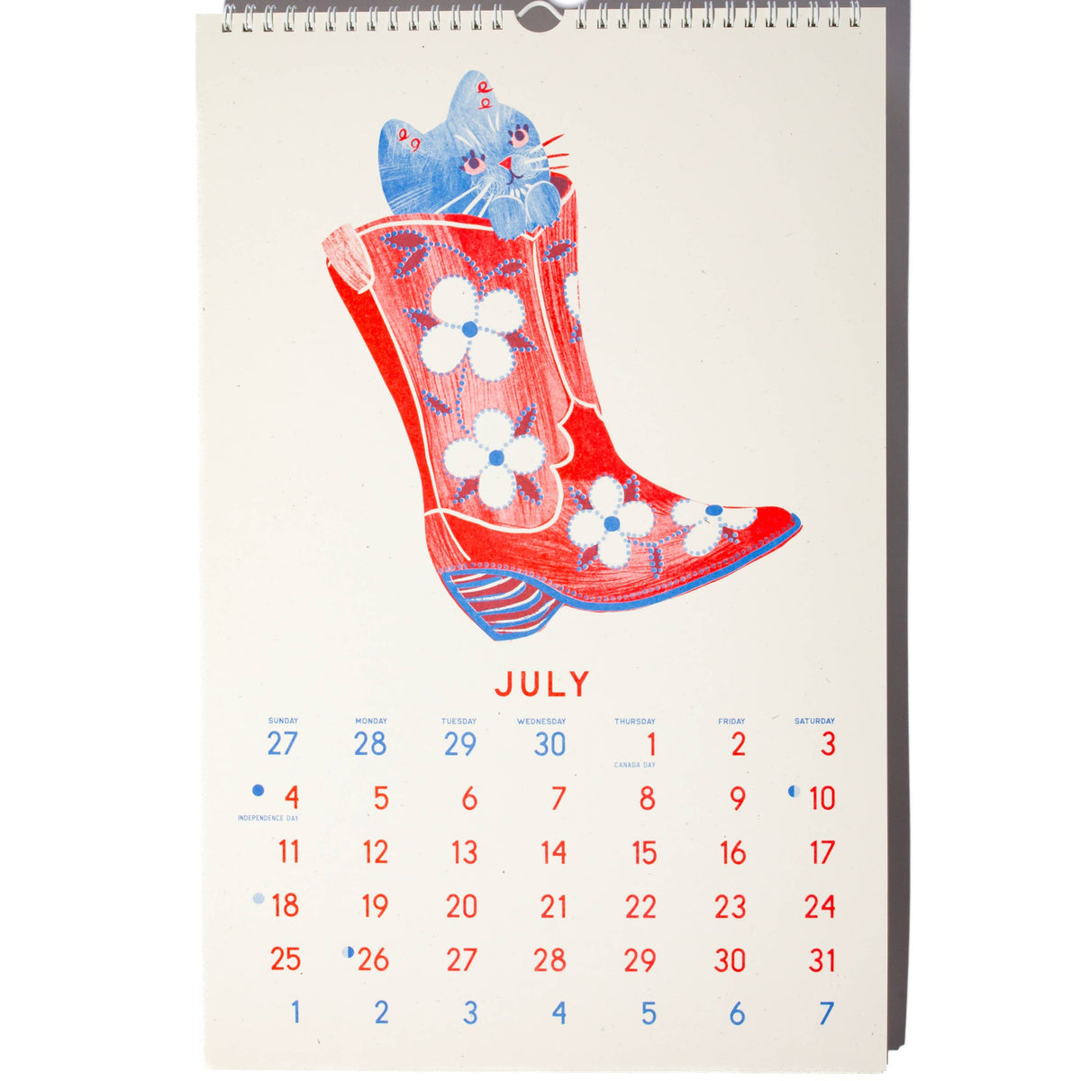 A spiral-bound July desk calendar featuring a whimsical illustration of a blue cat sitting inside a red cowboy boot with white flowers, on a white background.