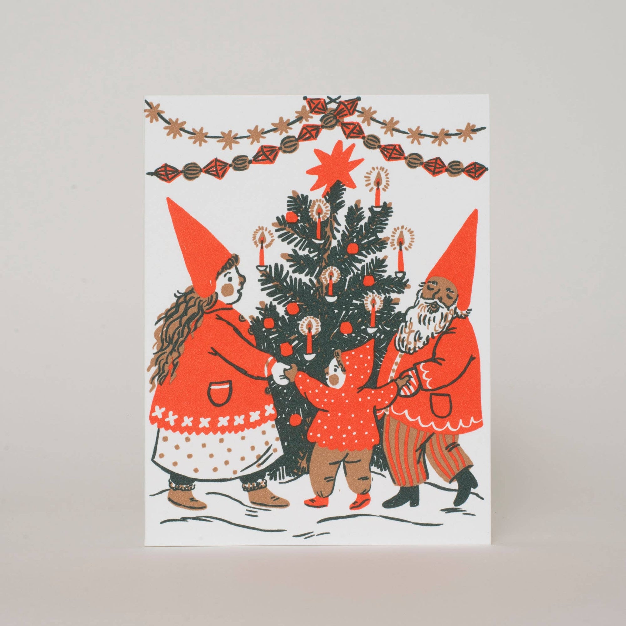 A vertical greeting card featuring a whimsical illustration of three Christmas gnomes decorating a tree, set against a plain light gray background.