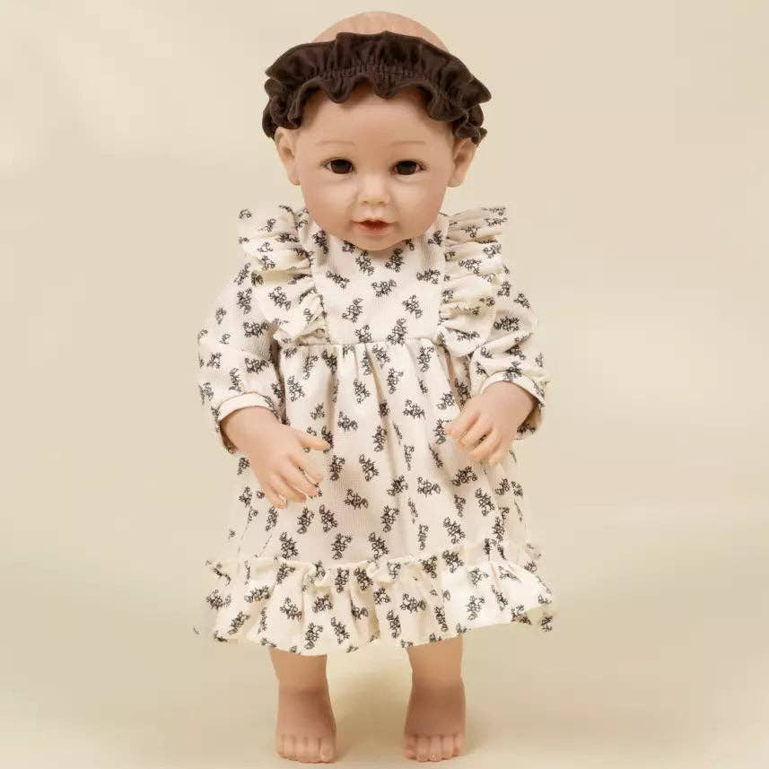 A vintage-style porcelain doll with dark brown curly hair, wearing a cream-colored dress with a small black floral pattern, standing against a solid beige background.