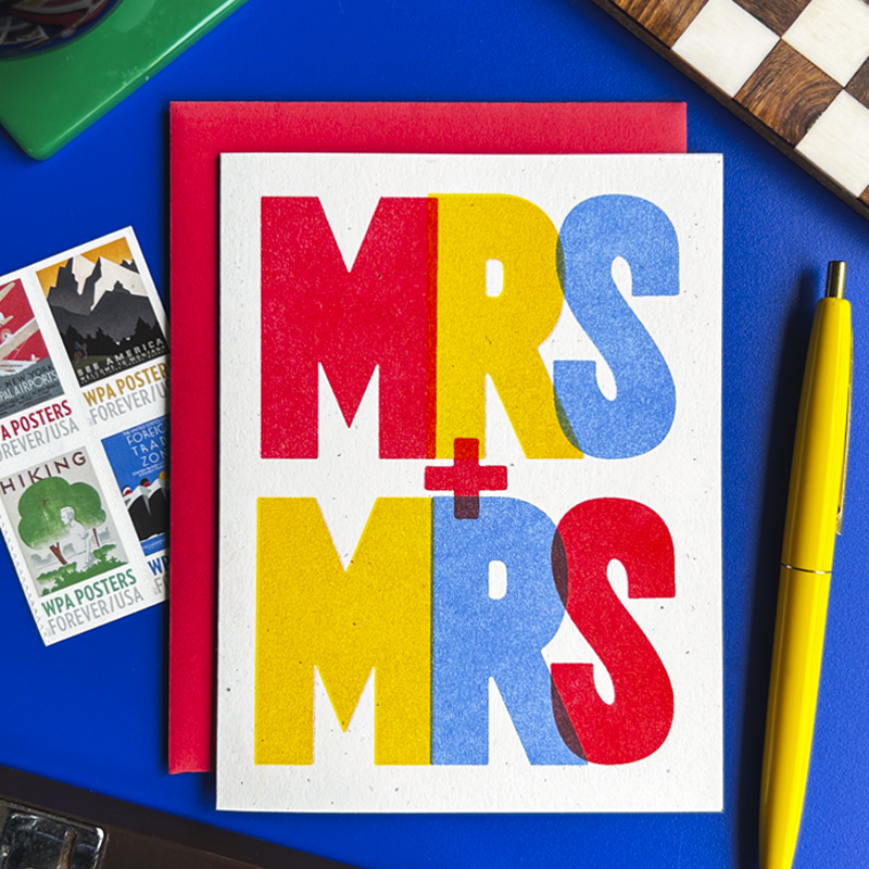 A square greeting card with a white background and bold colorful text reading 'MRS + MRS' in red, yellow, and blue, placed on top of a red envelope. The card is photographed on a blue desk with a yellow pen, vintage-style posters, and a chess piece visible around it.