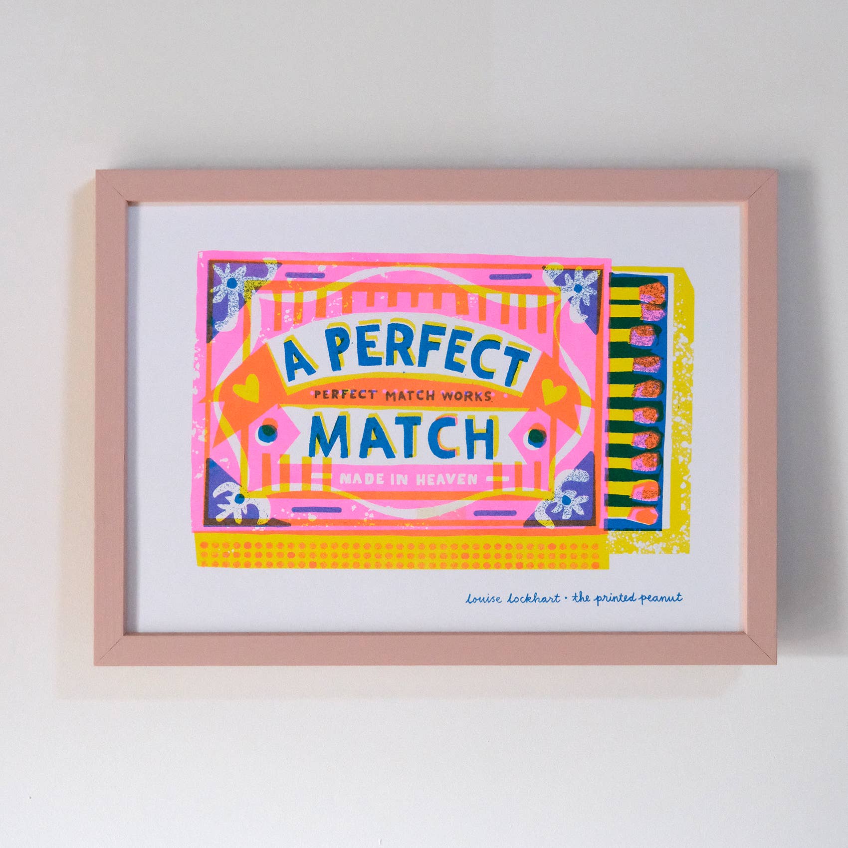 A framed art print featuring a colorful retro-style matchbox illustration with the text 'A PERFECT MATCH MADE IN HEAVEN' and a small signature at the bottom, displayed against a plain light gray wall.
