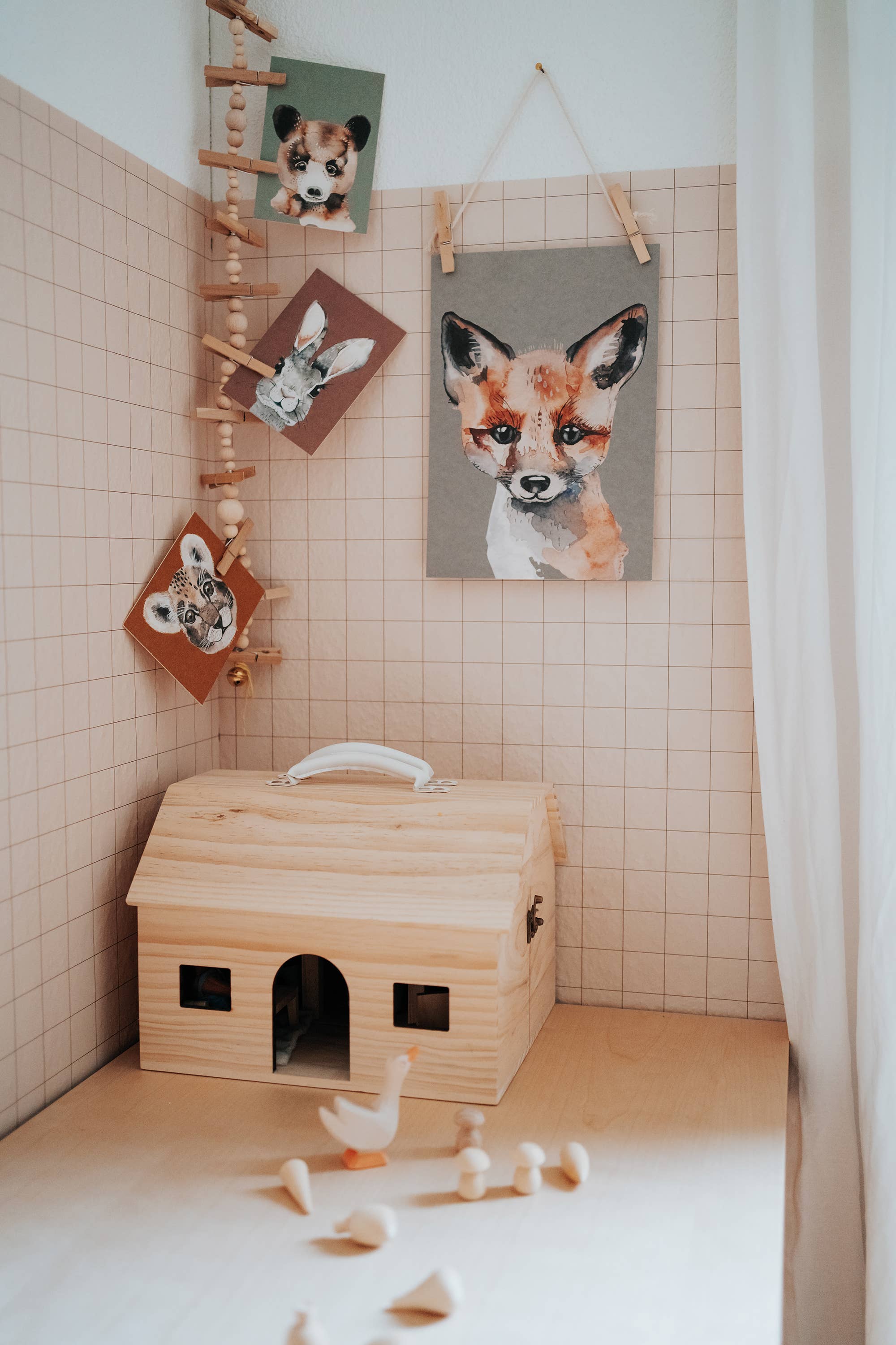 A wooden toy house with a small doghouse-shaped body and arched doorway, styled in a nursery-like room corner with pink tiled walls, hanging animal prints, a wooden ladder, and scattered wooden toy pieces on the floor.