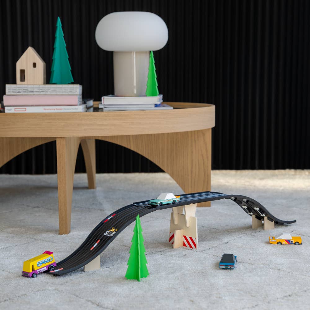 Toy car track set with small cars and trees on a wooden platform against a black background