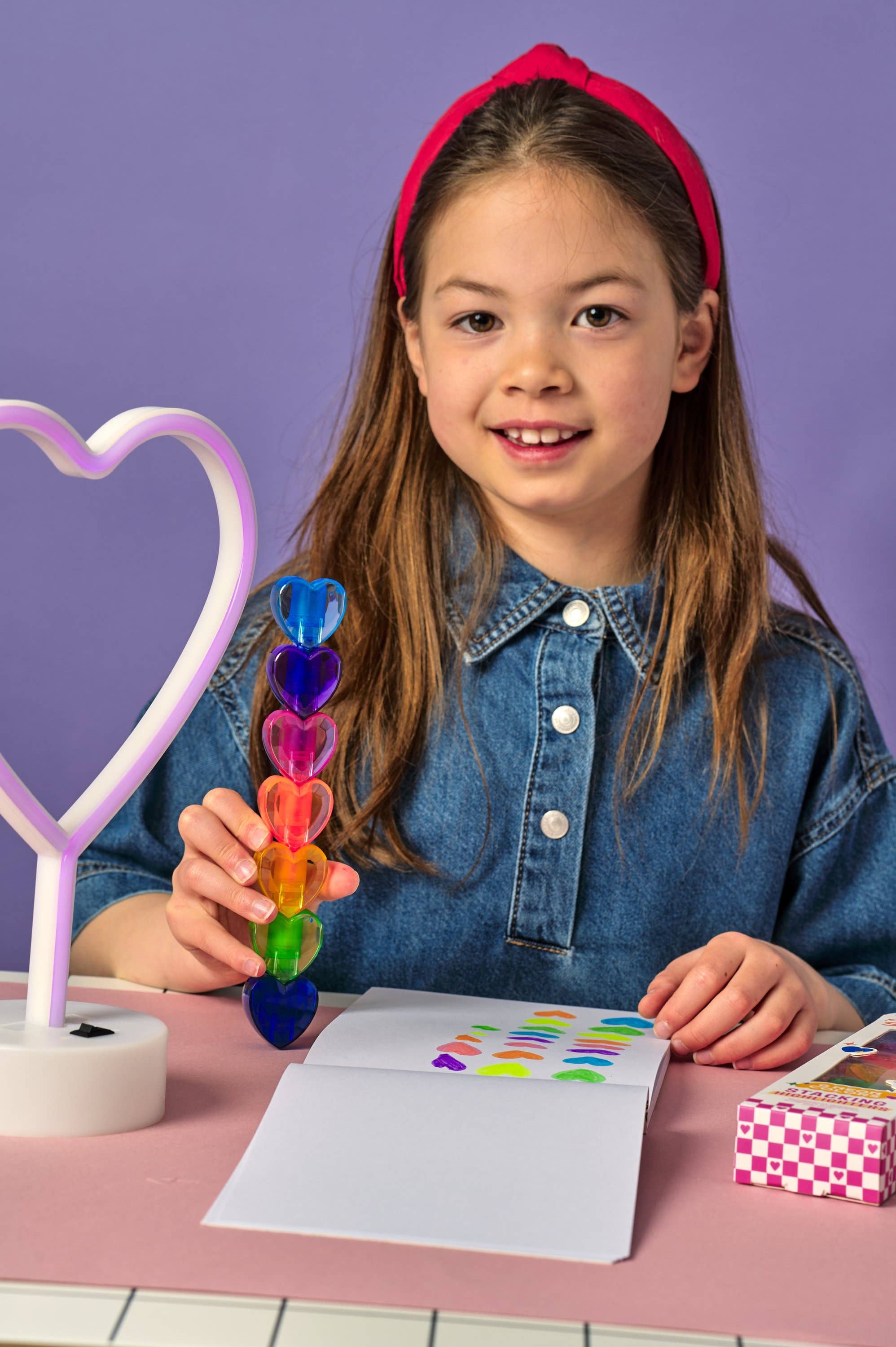 Young girl holding a colorful marker, sitting at a table with a heart-shaped lamp and paper.