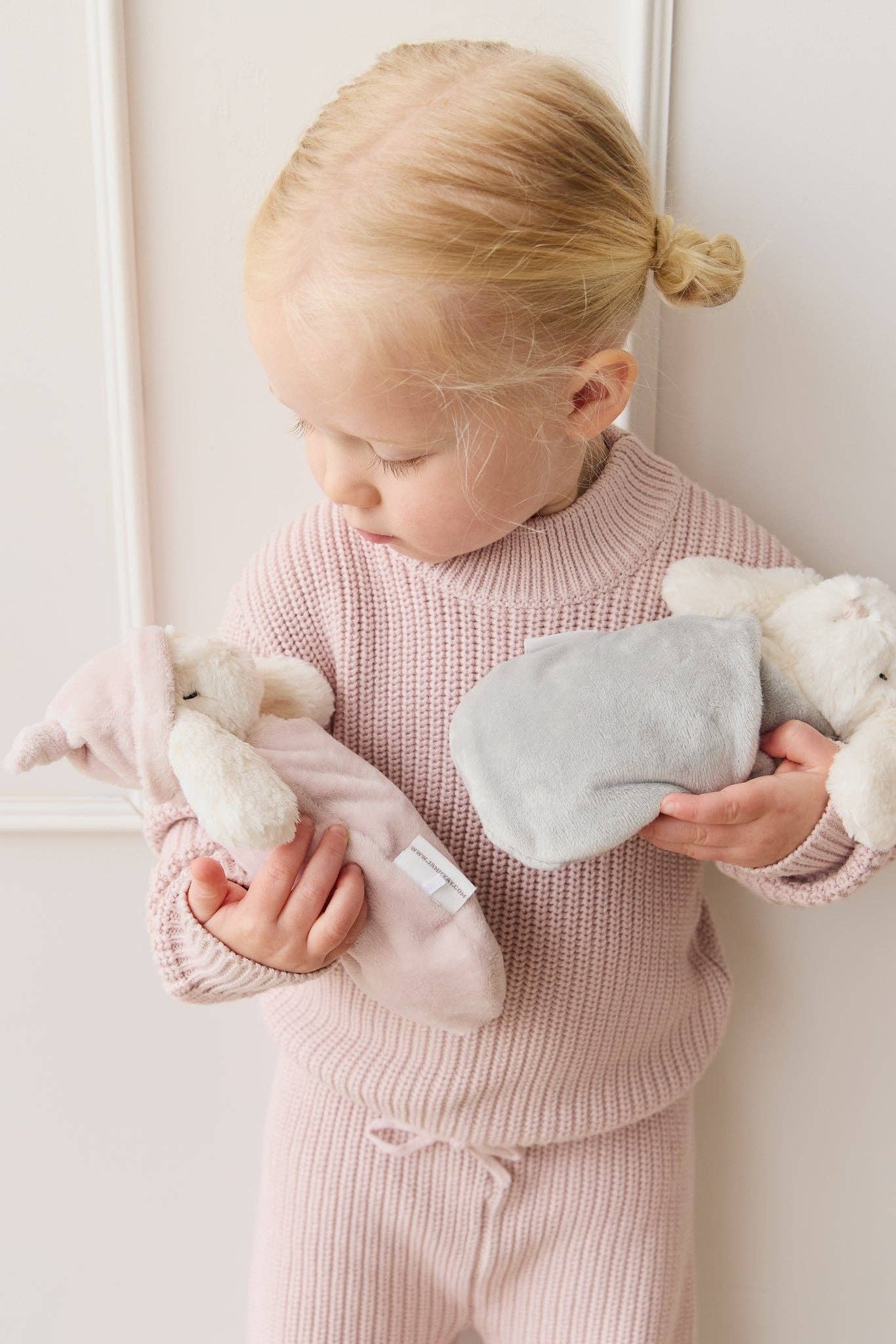 Child in pink outfit holding plush toys against a white wall