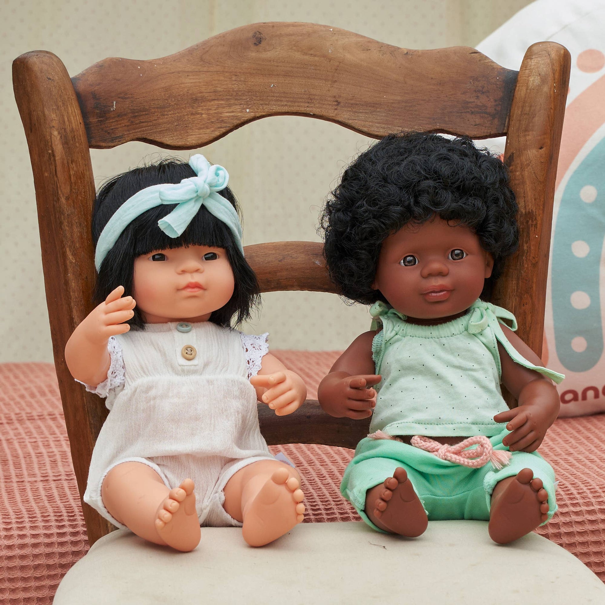 Two dolls sitting on a wooden chair with a soft pink blanket underneath.