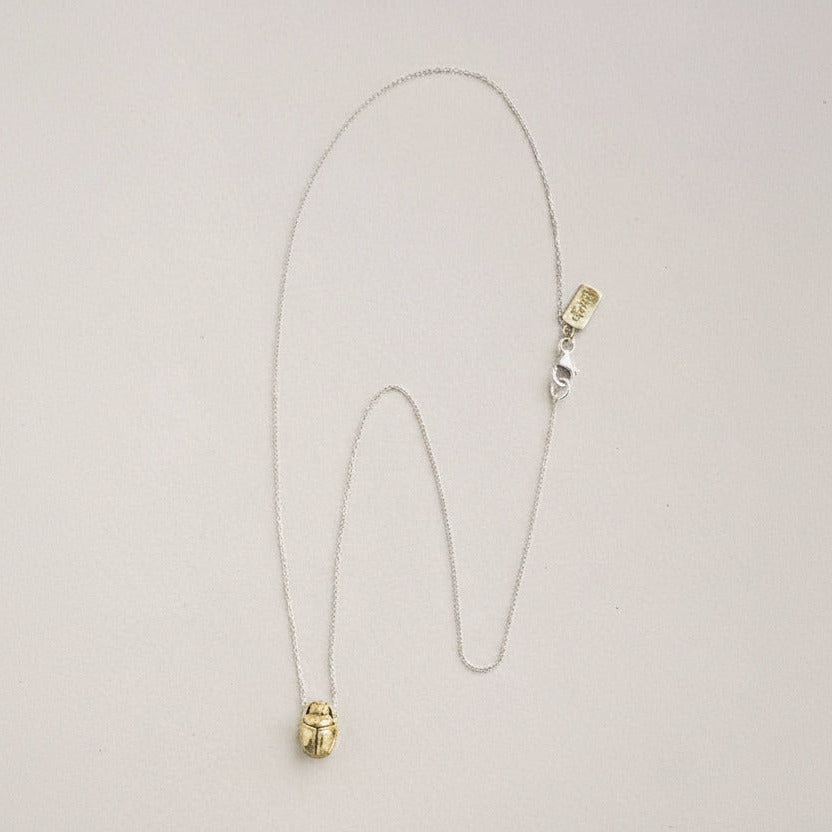 A delicate necklace with a small gold beetle pendant on a fine silver chain, displayed on a light beige background.