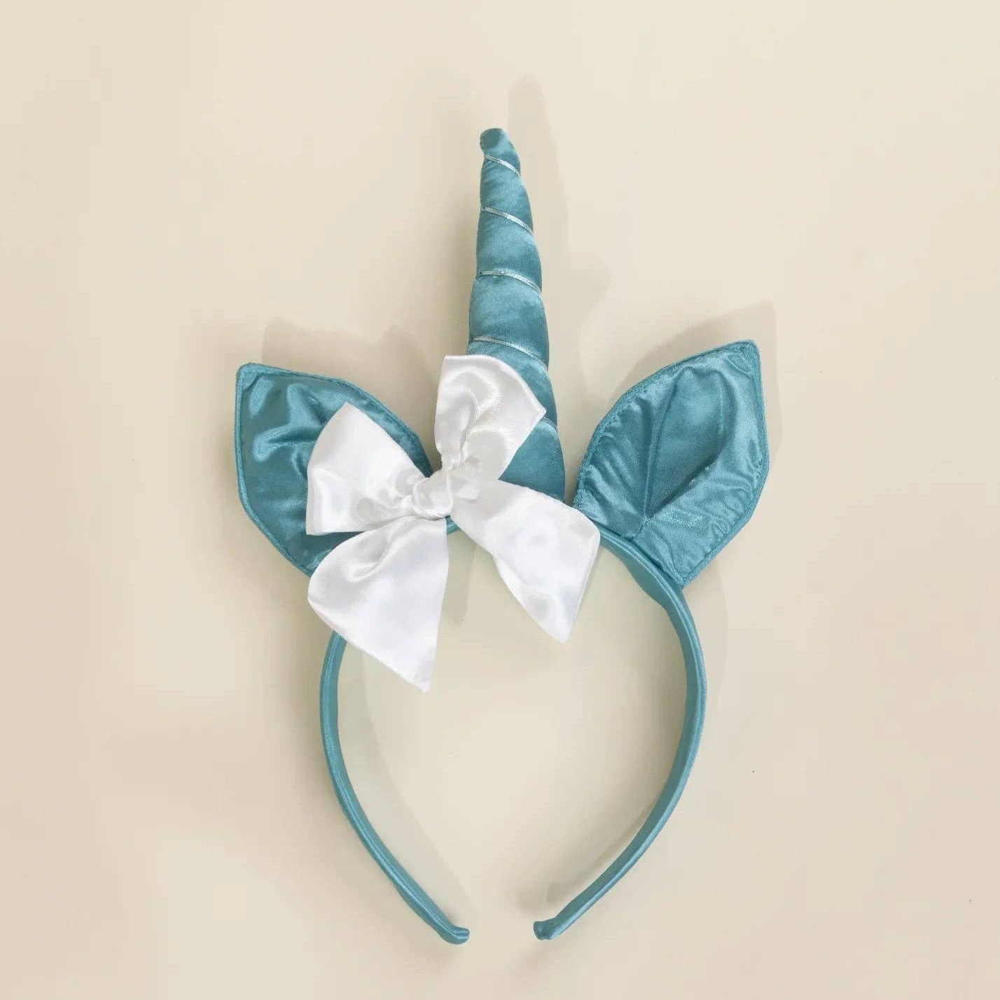 A unicorn headband featuring a teal blue satin horn, matching horn-ear silhouettes, and a white satin bow at the center, displayed on a solid beige background.