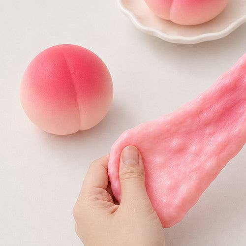 A hand stretches a pink peach-shaped squishy toy, with another peach-shaped toy resting on a white surface and a partial view of a matching toy on a small plate in the background.
