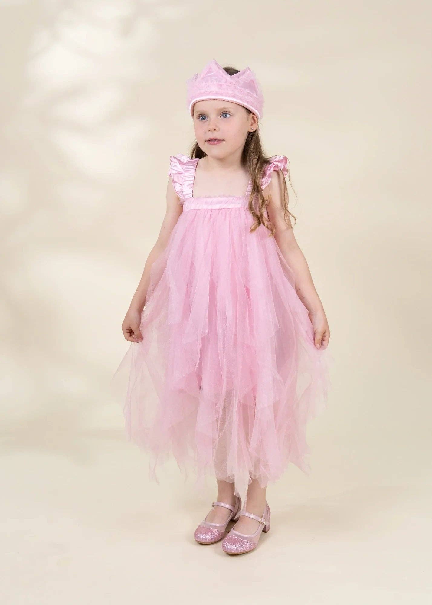 A young girl modeling a pink tulle dress costume with a matching pink headband and sparkly pink shoes, standing against a warm beige studio background.