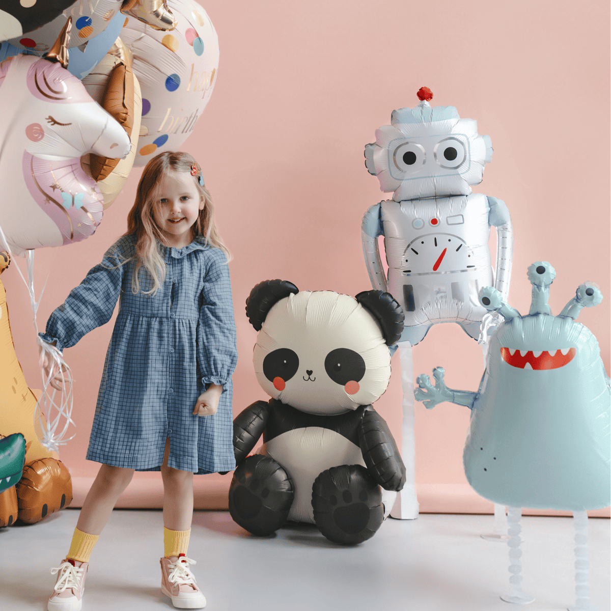 Child in a blue dress standing next to inflatable panda, robot, and monster toys against a pink background.