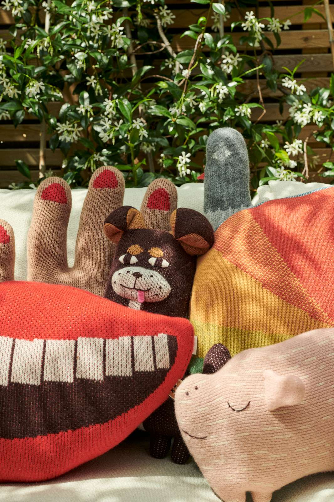 A group of colorful, novelty knit animal-shaped pillows arranged together on a light-colored surface with a garden background. The pillows include a red and brown toothy grin, a brown dog, a yellow and orange striped figure, a gray finger, and a pink pig.