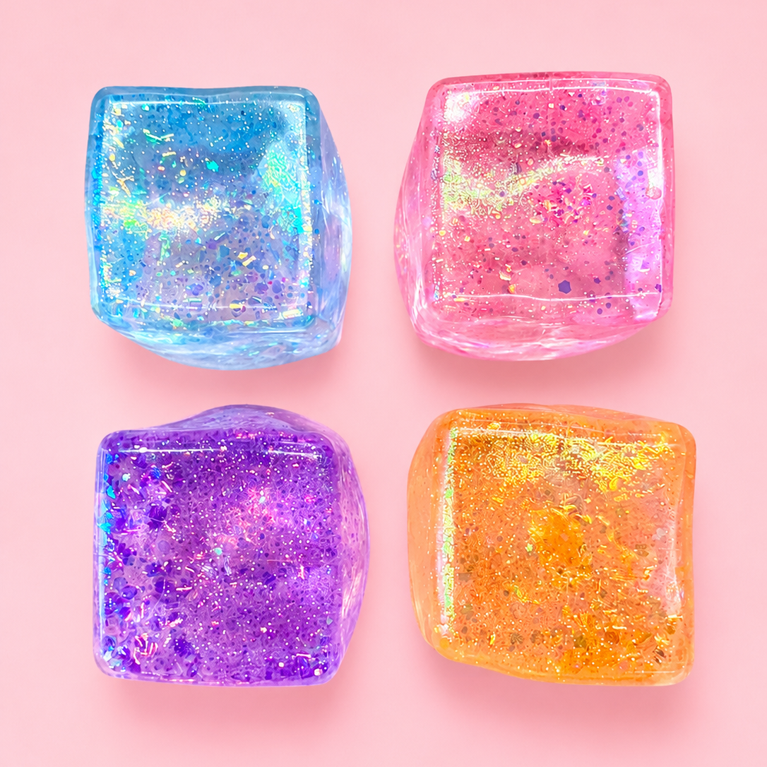 Four translucent glitter ice cube charms in blue, pink, purple, and orange, arranged in a 2x2 grid on a solid pink background.