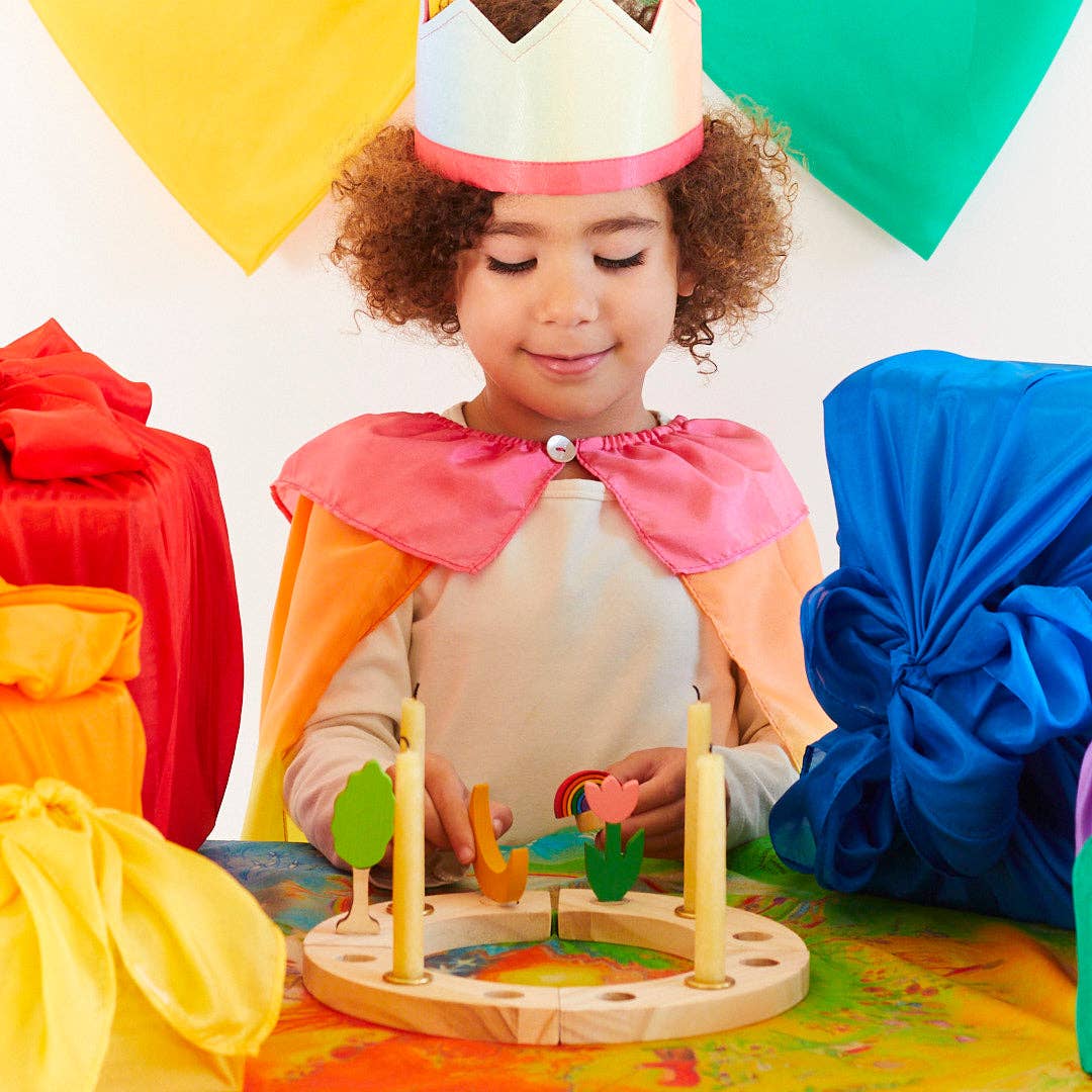 Child in colorful costume playing with a wooden toy, surrounded by colorful balloons and costumes.