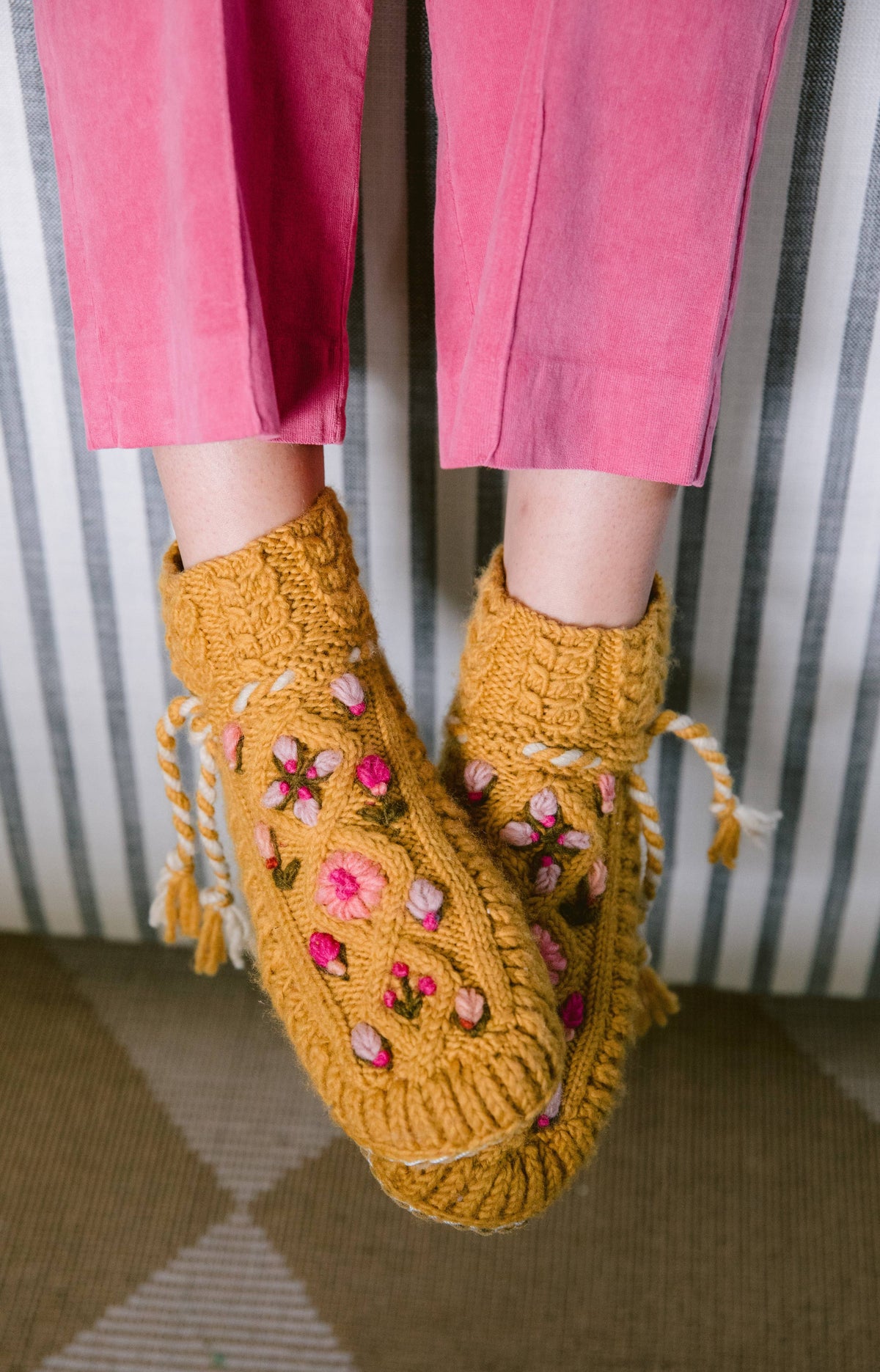 Yellow knitted slippers with floral patterns worn by a person in pink pants against a striped background.