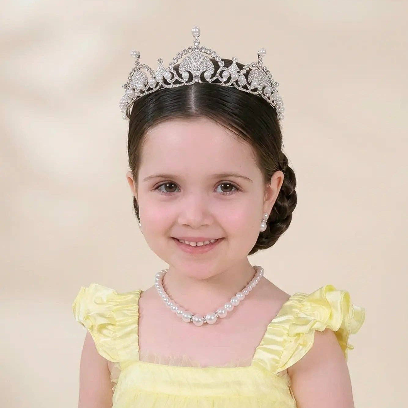 A young girl with dark hair styled in a braid, wearing a silver tiara, a pearl necklace, and a pale yellow dress with ruffled shoulders, set against a soft beige background.