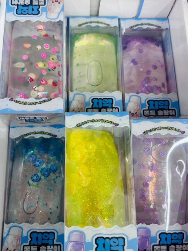 Six packaged translucent phone cases arranged in a grid inside a box. The cases contain colorful glitter and charms, including pink, green, purple, blue, yellow, and iridescent designs.