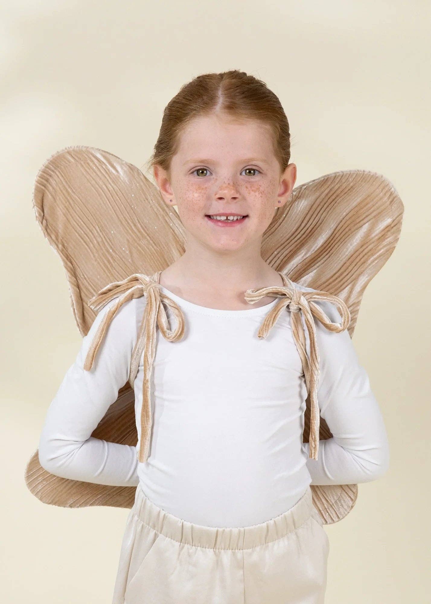 A young girl with freckles wearing a white long-sleeve bodysuit and beige angel wings with matching tie straps, posing against a solid light beige background.