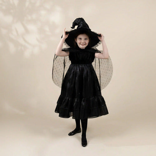 A young girl modeling a black witch costume dress with a matching pointed witch hat, sheer polka-dot wings, and black tights against a warm beige studio background.
